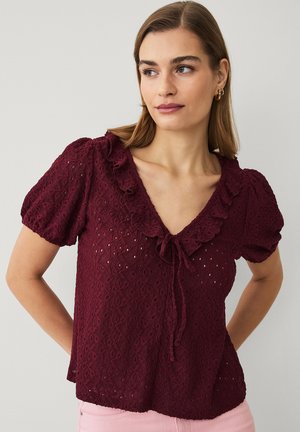 Frau trägt eine burgunderrote Spitzenbluse mit Puffärmeln und gerafftem V-Ausschnitt, vorne gebunden, kombiniert mit hellrosa Hose.