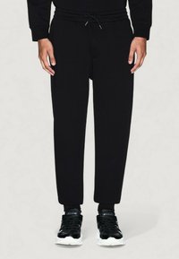 Modelo masculino vistiendo pantalones de chándal negros con cordones y zapatillas negras con suelas blancas, de pie frente a un fondo claro y liso.