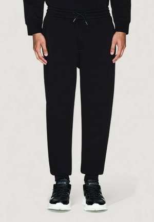 Modello maschile che indossa pantaloni jogger neri con cordoncino e sneakers nere con suola bianca, in piedi davanti a uno sfondo chiaro e uniforme.
