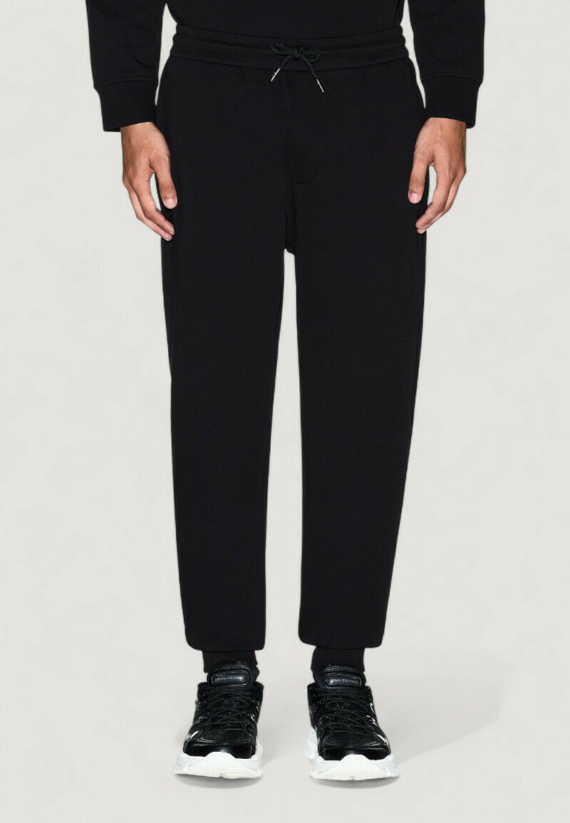 Modelo masculino vistiendo pantalones de chándal negros con cordones y zapatillas negras con suelas blancas, de pie frente a un fondo claro y liso.