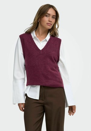 Femme portant une chemise blanche à manches longues avec de larges poignets, un pull sans manches en maille violet à col en V et un pantalon marron sur un fond uni.