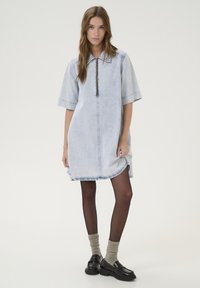 Robe chemise en denim bleu clair, à manches courtes, fermeture éclair à l'avant, fentes sur les côtés et ourlet arrondi. Associée à des bas foncés et des mocassins.
