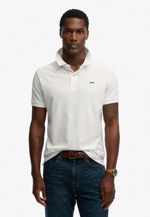 Witte katoenen poloshirt met een omgeslagen kraag en een knopenbies met twee knopen. Draagt blauw spijkerbroek en een bruine gevlochten riem. Minimale merkopdruk op de borst.