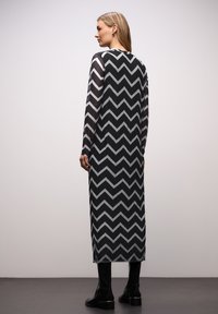 Černobílé dlouhé šaty s chevronovým vzorem, dlouhými rukávy a plisovanou texturou, doplněné černými kotníkovými botami.