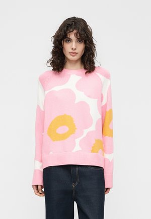 Kvinde med krøllet hår iført en lyserød sweater med store hvide og gule blomsterprint og mørkeblå jeans, stående mod en ensfarvet baggrund.