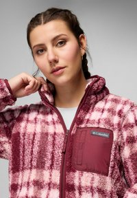 Giovane donna con capelli intrecciati che indossa una giacca di pile a quadri marrone e rosa, aggiustando il colletto su uno sfondo grigio semplice.