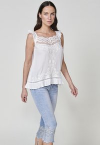 Top sans manches blanc avec des détails en dentelle et des bretelles volantes, associé à un jean cropped bleu clair avec des ourlets brodés.