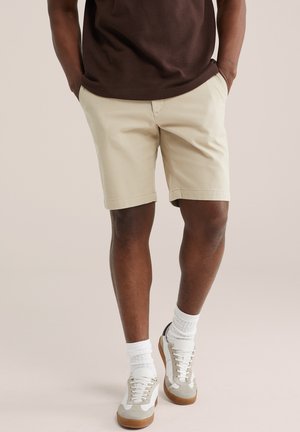 Mann trägt beige Shorts, weiße Sportsocken, beige-weiße Turnschuhe und ein braunes Hemd, die Hände in den Taschen, steht vor einem schlichten Hintergrund.