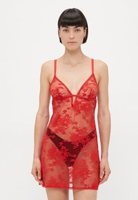 Woman wearing sheer red lace lingerie slip with floral patterns and thin shoulder straps, standing against white background.