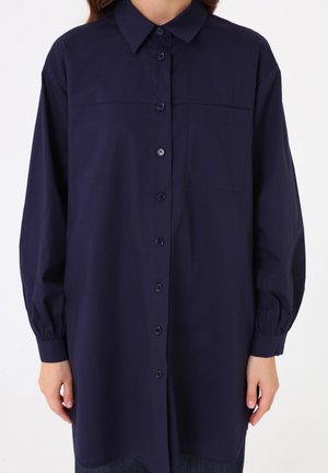Femme portant une chemise ample bleu marine à manches longues avec col et poche poitrine, sur un fond clair uni.