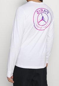 Vit långärmad t-shirt med en rund grafik på baksidan som visar "PARIS" och en basketbollssiluett i rosa och lila nyanser.