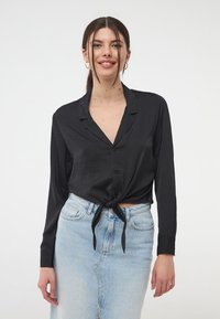 Camicia nera a maniche lunghe con bottoni e design con nodo frontale, realizzata in un materiale liscio; abbinata a una gonna di jeans azzurri.