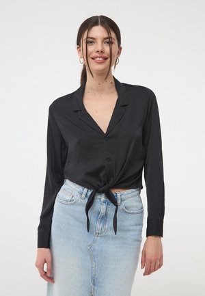 Camicia nera a maniche lunghe con bottoni e design con nodo frontale, realizzata in un materiale liscio; abbinata a una gonna di jeans azzurri.