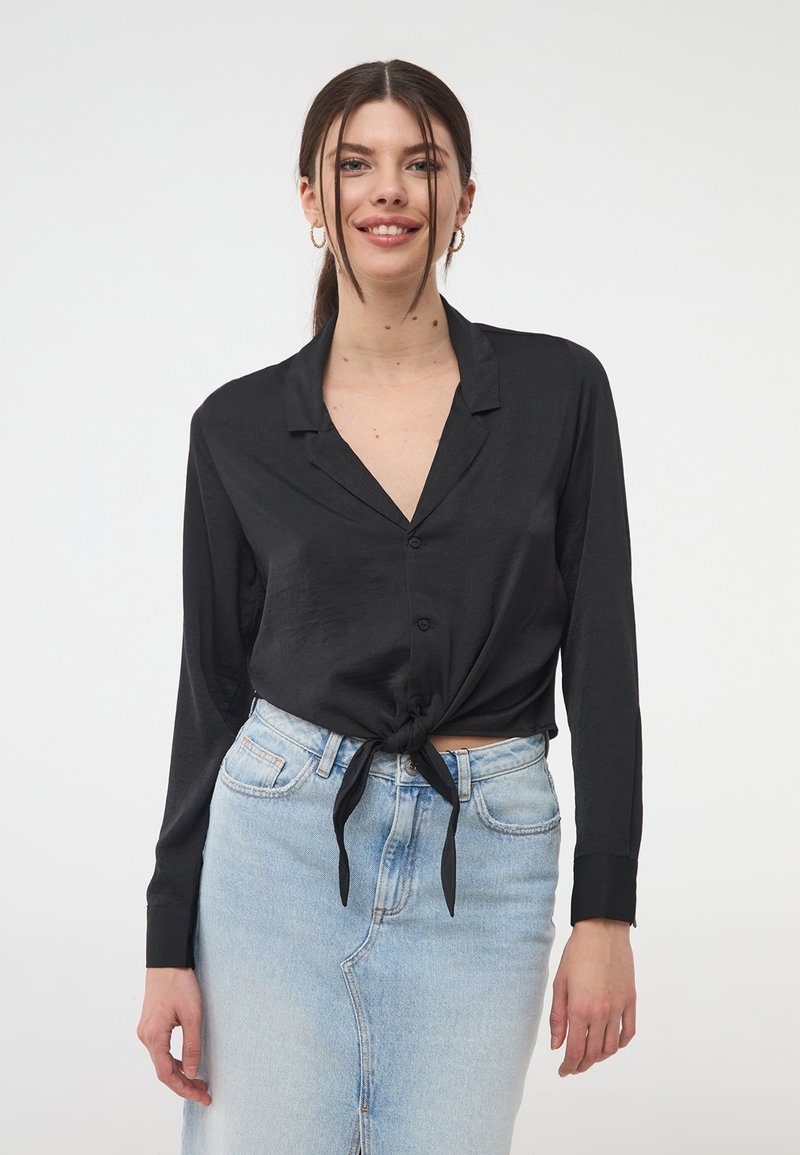 Camicia nera a maniche lunghe con bottoni e design con nodo frontale, realizzata in un materiale liscio; abbinata a una gonna di jeans azzurri.