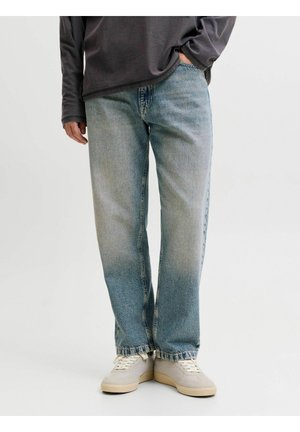 Personne portant un jean droit bleu clair, des baskets beiges et une chemise à manches longues foncée, une main dans la poche.