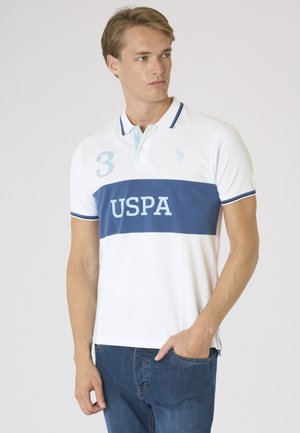 Giovane uomo che indossa una polo bianca con una striscia orizzontale blu e la scritta "USPA", abbinata a jeans blu, in piedi su uno sfondo semplice.