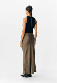 Donna con lunghi capelli intrecciati che indossa un top senza maniche nero, una gonna maxi marrone e scarpe flat nere, in piedi su uno sfondo bianco uniforme.