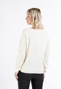 Crème kleurige trui met een relaxte pasvorm, voorzien van gouden hartaccenten op de mouwen, ribgebreide zoom en een zachte textuur. Gemanipuleerd met zwarte jeans.