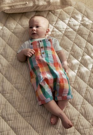 Bébé allongé sur une couverture matelassée beige, portant une salopette à carreaux colorée et une chemise blanche à manches courtes, regardant légèrement sur le côté.