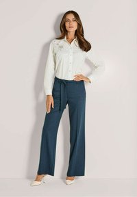 Vrouw in een witte geborduurde blouse met knopen, marineblauwe wijdvallende broek met een strik, en witte puntige hakken, staand tegen een effen muur.