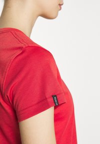 Side view of person wearing a red short-sleeve shirt with "4X FASTER" black tag on the sleeve against a white background.