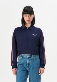 Cropped navy sweatshirt with a zip front, ribbed hem, and collar. Features white and maroon stripes along the sleeves and "GAP NYC" logo.