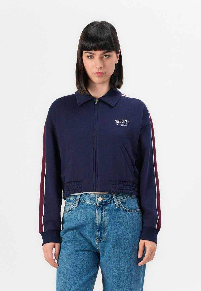 Cropped navy sweatshirt with a zip front, ribbed hem, and collar. Features white and maroon stripes along the sleeves and "GAP NYC" logo.