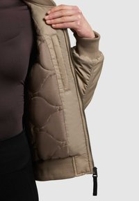 Beige bomber bunda so prešívaným interiérom, rebrovanými manžetami a zipsom. Tkanina má hladkú textúru a jemný lesk.