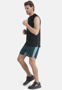 Svart ärmlös topp, teal och gröna shorts med sidopaneler, och svarta löparskor med röda accenter. Positionerad i en löpställning.