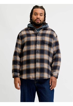 Homme aux cheveux tressés portant une chemise à carreaux marron et noire boutonnée par-dessus un sweat à capuche sombre et un jean bleu sur un fond uni.
