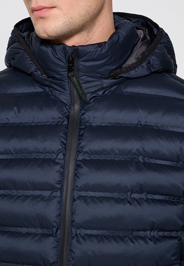 Granatowa kurtka puchowa z zapięciem na zamek, kapturem i pikowaną fakturą. Posiada czarne akcenty oraz gładkie, błyszczące wykończenie.