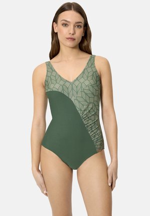 Mujer con un traje de baño enterizo verde con un estampado de hojas en la parte superior y color sólido en la parte inferior, posando contra un fondo blanco.