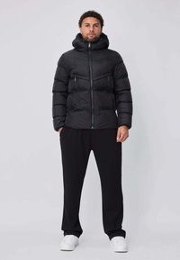 Doudoune noire à capuche, avec une fermeture éclair frontale et des poches latérales, associée à un pantalon noir et des baskets blanches.