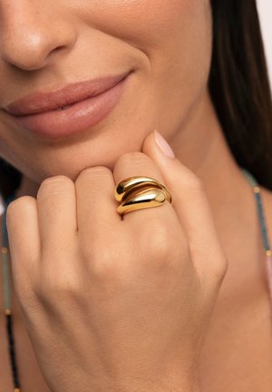 Anillo de oro con dos bandas entrelazadas, lisas y pulidas. La mano tiene una textura natural de piel y una laca de uñas de tono neutro.