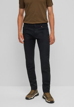 Hombre con pantalones negros ajustados, camisa marrón de manga corta y zapatos de senderismo marrones de pie frente a un fondo gris liso.
