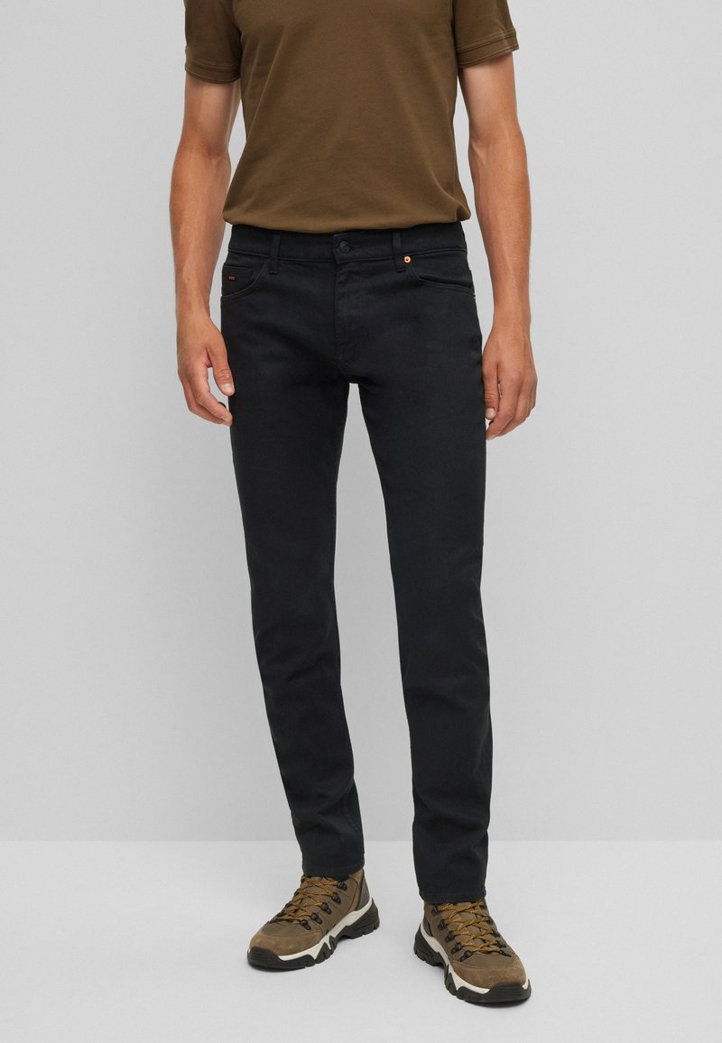 Homme portant un pantalon noir slim, une chemise marron à manches courtes et des chaussures de randonnée marron, debout devant un fond gris uni.