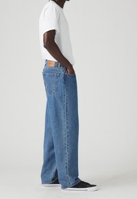 Person som bär lösa blå jeans, vit t-shirt och svarta sneakers, står sidledes med händerna i fickorna mot en enkel bakgrund.