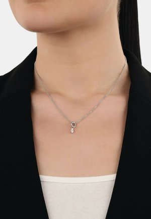 Collana d'argento con design a catena, caratterizzata da un pendente rotondo e un accento a goccia. Indossata sopra una maglietta chiara contro una giacca nera.