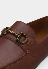 Καφέ δερμάτινο μοκασίνι με χρυσό μεταλλικό διακοσμητικό σχήματος αλόγου πάνω από το επάνω μέρος και εμφανείς ραφές κατά μήκος των άκρων.