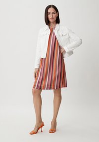 Veste en denim blanche portée sur une robe à rayures multicolores avec des teintes de rose, d'orange et de blanc, accompagnée de sandales à talons hauts orange.