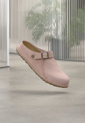 Zueco deslizante de ante rosa claro con correa ajustable con hebilla y suela beige texturizada, mostrado en el aire sobre suelo de hormigón en interiores.