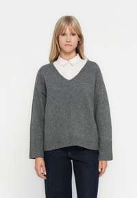 Pull en maille gris avec un col en V, épaules tombantes, et des poignets et un ourlet côtelés. Associé à une chemise à col claire en dessous.