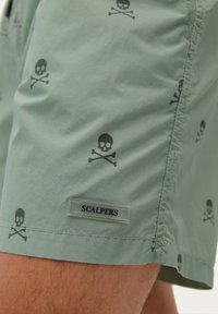 Shorts de bain verts en tissu léger, présentant un motif répété de crânes et os croisés, avec une étiquette "SCALPERS".