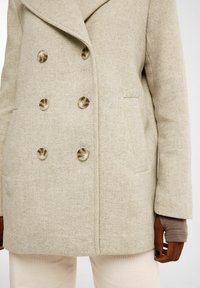 Beige takki, jossa on kaksinkertainen nappikiinnitys ja suuret metalliset napit, herringbone-kuviointi ja sivutaskut. Sujuva, muotoiltu design.