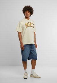 Cremefarvet bomulds T-shirt med et flerfarvet logo print, parret med blå denim shorts. Modellen har hvide sneakers og ankelsokker på.