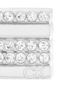 Hopeinen metallinen suorakaiteen muotoinen kappale, jossa on riveittäin kirkasta rhinestone-koristeita. Yhdellä puolella on kaiverretut kirjaimet "CK". Pintakuvio on sileä ja kiiltävä.