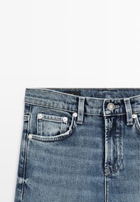 Niebieskie dżinsy z denimu, odcinek w talii z guzikiem, zamkiem błyskawicznym, szlufkami na pasek oraz przednimi i małą kieszonką, prezentowane na białym tle.