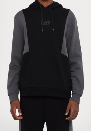 Man draagt een zwarte en donkergrijze color-blocked hoodie met lange mouwen en een logo aan de voorkant, gecentreerd op de borst.