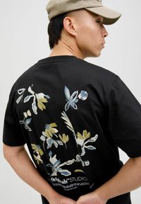 Schwarzes Baumwoll-T-Shirt mit floralem Druck auf der Rückseite in Blau, Gelb und Weiß. Beinhaltet Markentext am unteren Rand.