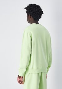 Sudadera verde claro con cuello redondo y mangas largas. Textura suave, puños y cintura acanalados. Vista desde la parte trasera.